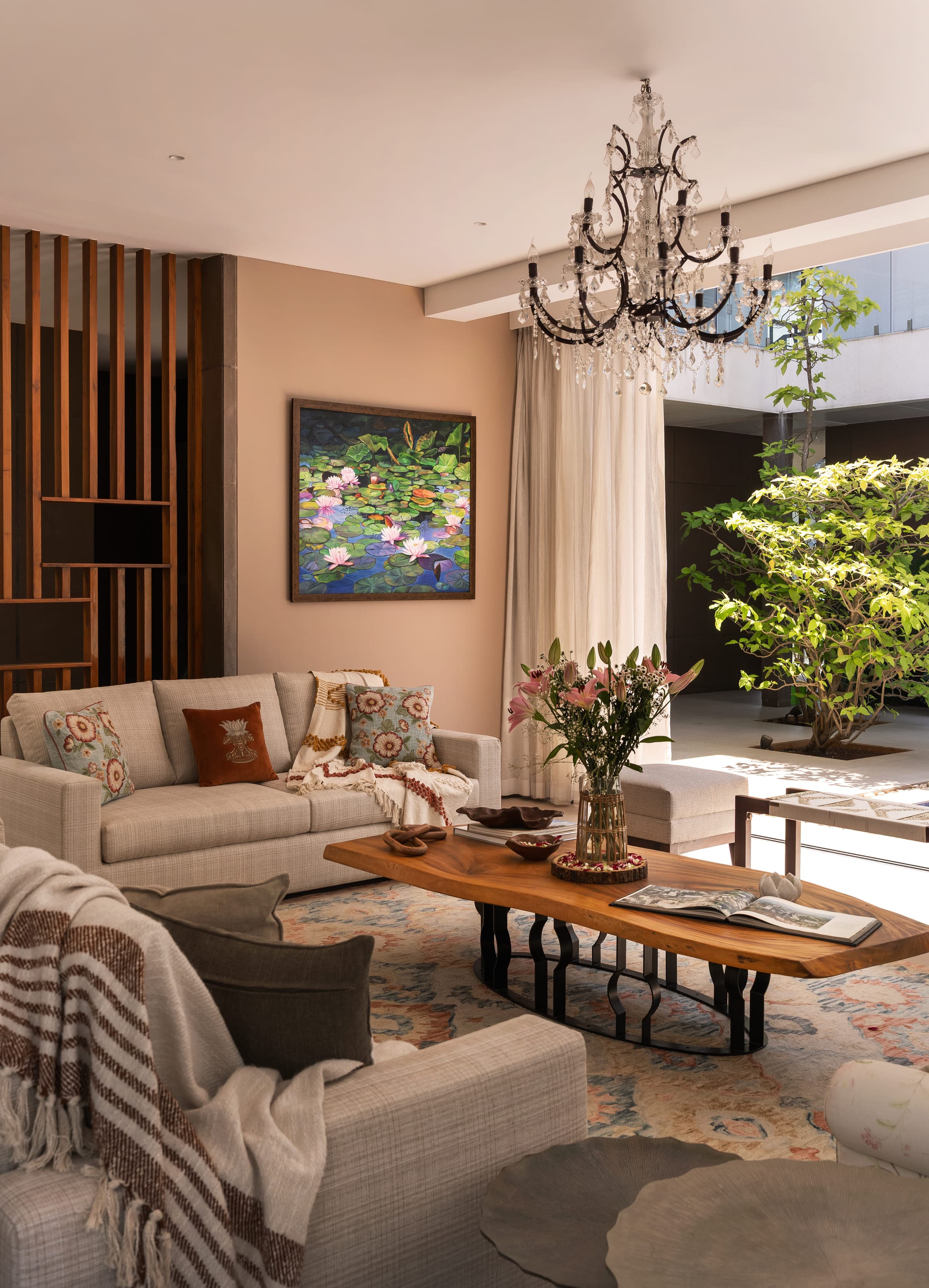 The living and dining pavilion of the house in Alibaug: linen seating, a solid-log coffee table, a lotus painting on the far wall and a crystal chandelier overhead.