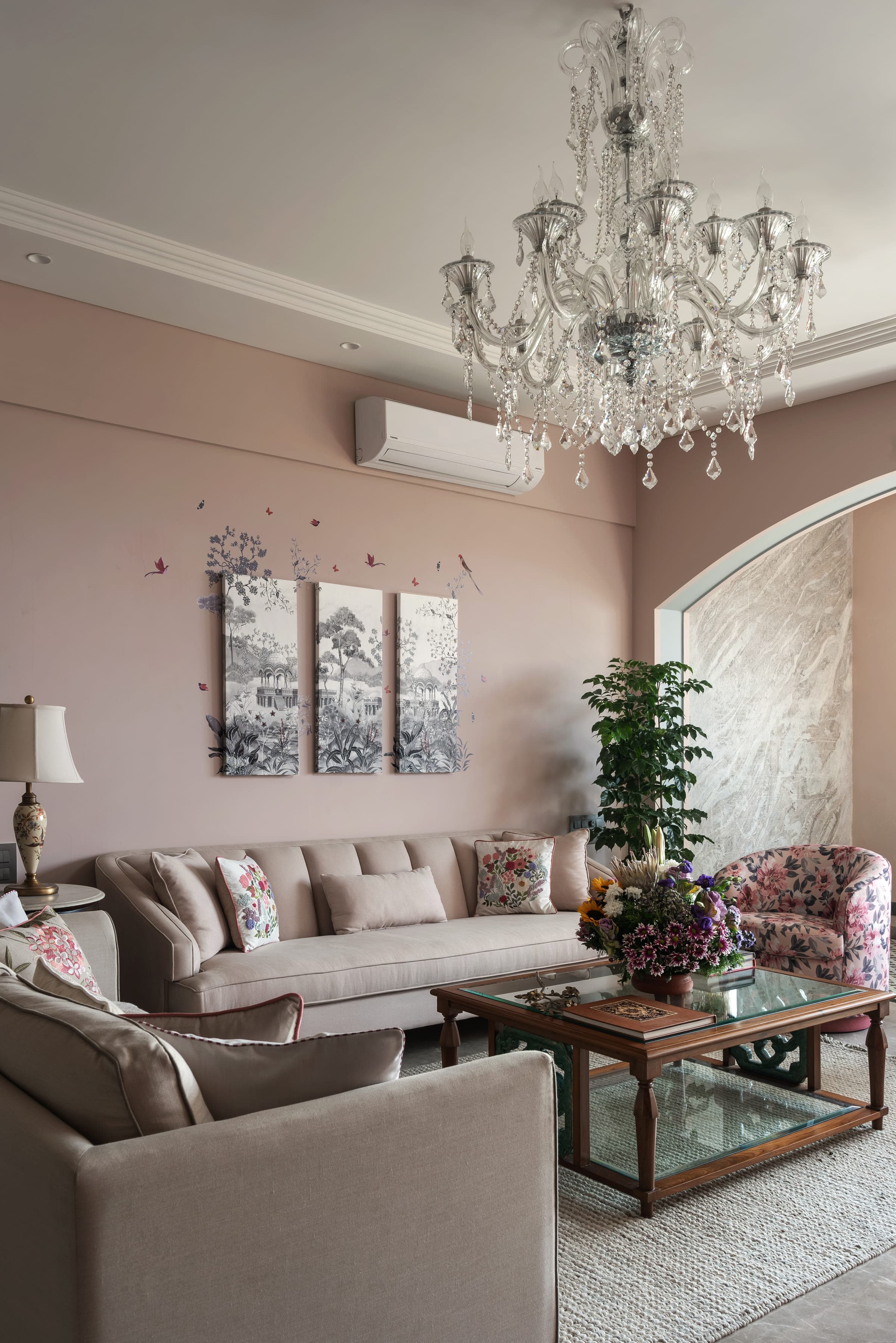 A living room at Family Home in Chowpatty: a long pale sofa beneath a triptych of botanical prints, a crystal chandelier overhead and a tall plant in the corner.